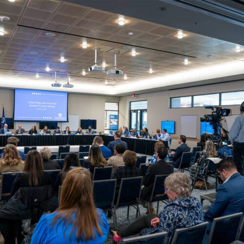 A wide-angle view of a group of people in a meeting in a large, modern conference room. some are seated along U-shaped table at the front, facing the rest. A large projection screen displays in the background